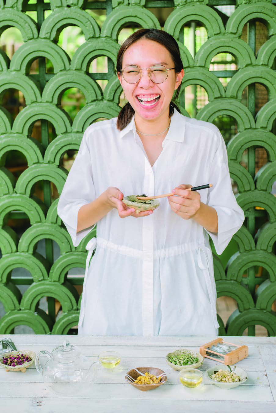 ผู้ดูแลบ้านใบยิ้มอยู่หน้าผนังบล็อกช่องลมสีเขียว พร้อมถ้วยและวัตถุดิบชา